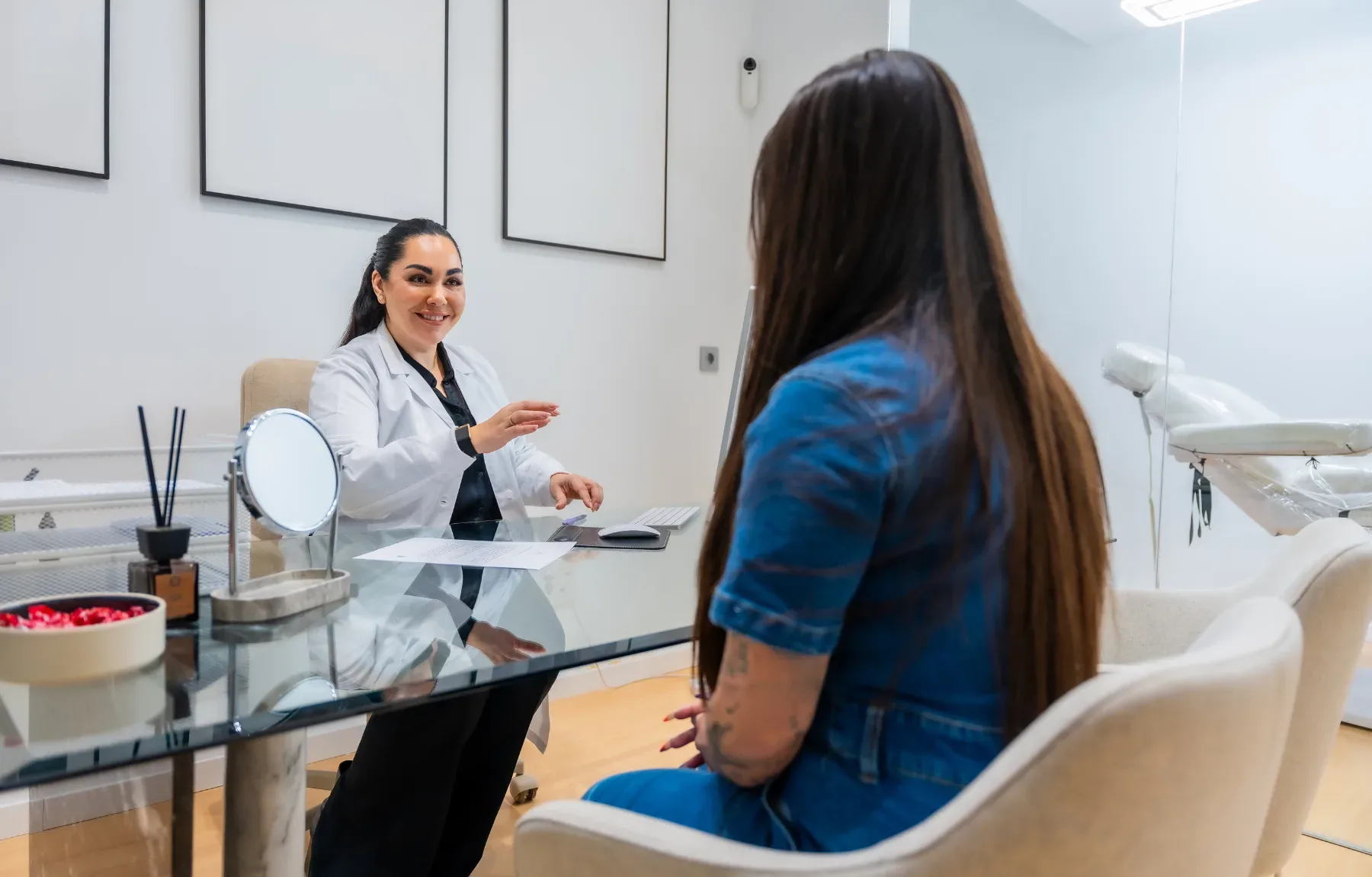A smiling woman in a white lab coat and black tie sits behind a glass desk, gesturing while talking to a client with long dar