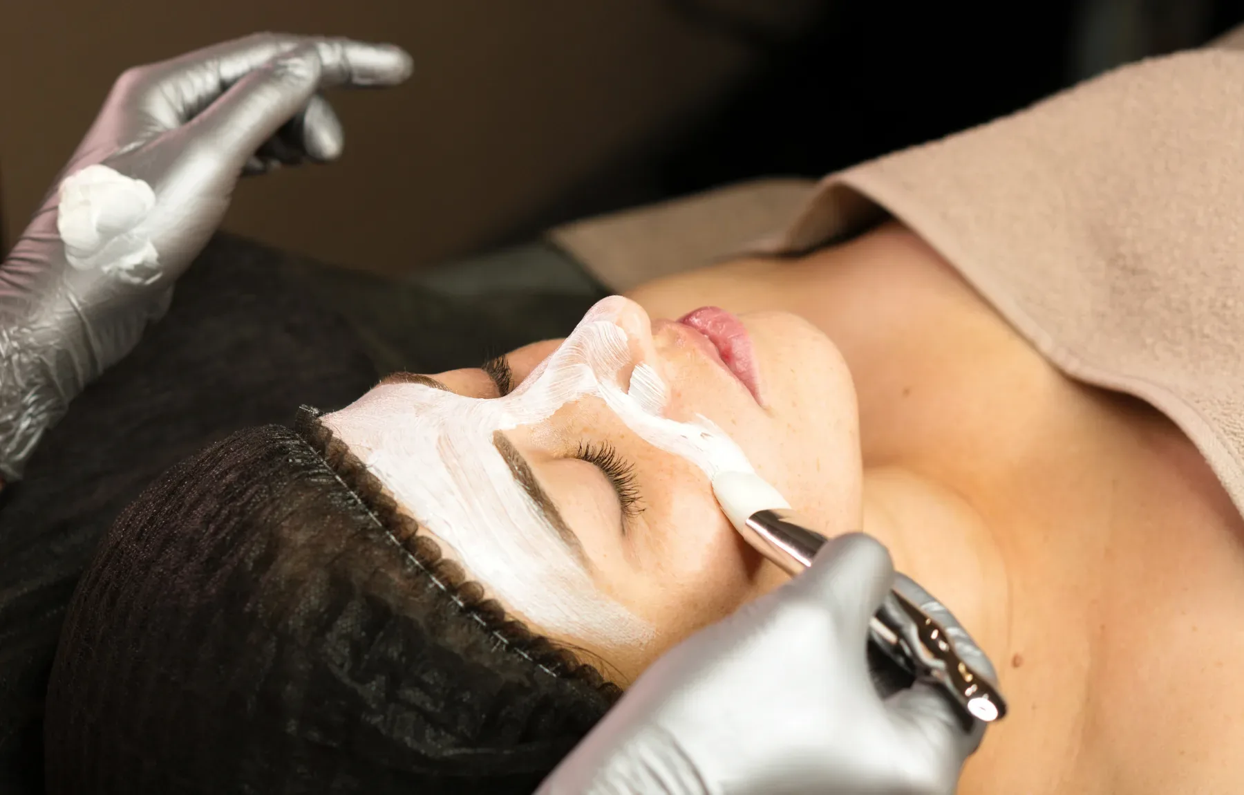 A woman with a white towel wrapped around her head lies with her eyes closed while a gloved hand brushes a peach-toned mask o